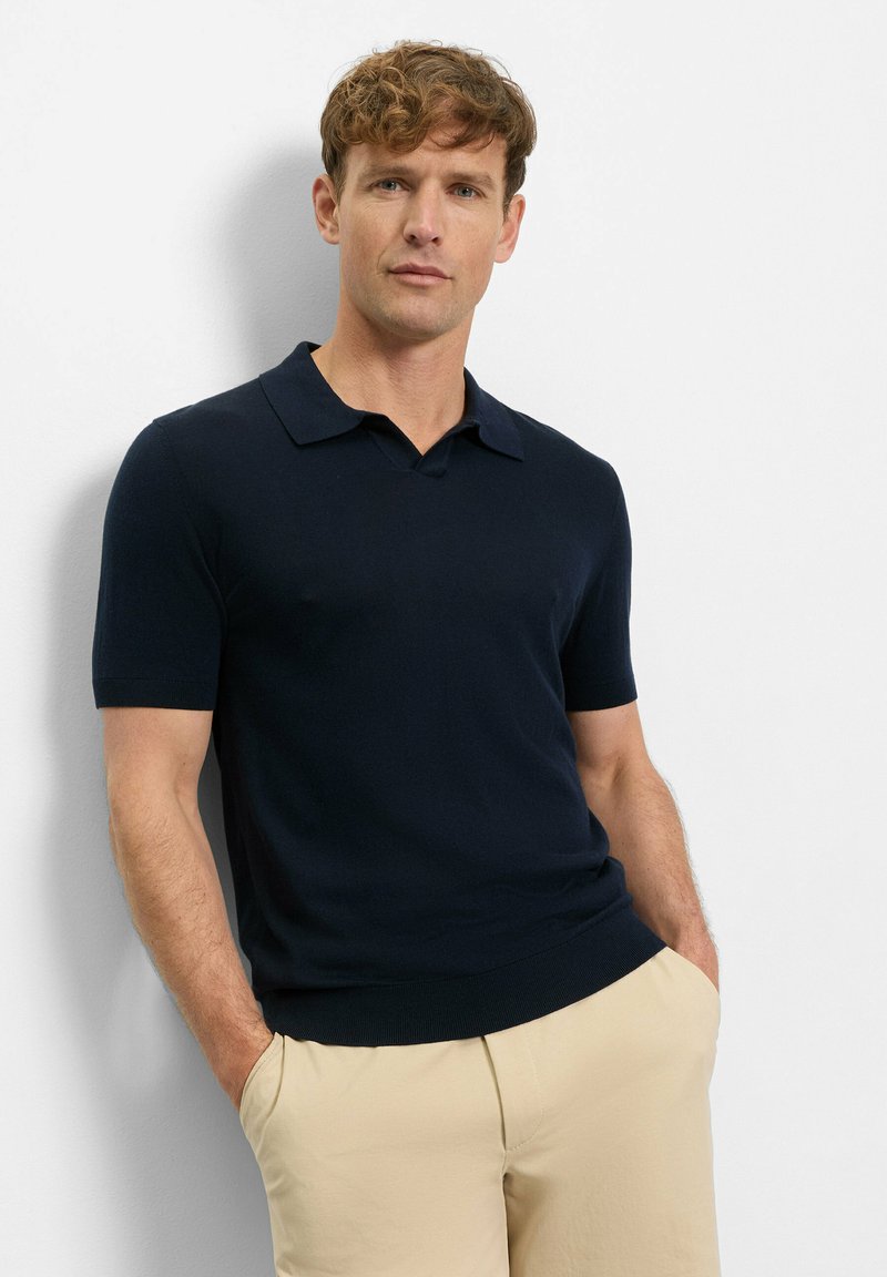 Mann mit kurzen lockigen Haaren, der ein marineblaues Polohemd mit kurzen Ärmeln und beige Hose trägt, steht mit den Händen in den Taschen vor einer weißen Wand.