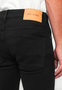 Acercamiento de una persona que lleva vaqueros negros con un parche de cuero marrón claro etiquetado como "ONLY & SONS" encima del bolsillo trasero derecho.