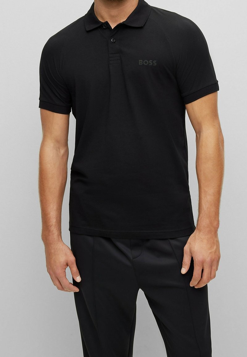 Polo shirt noir à manches courtes avec col, arborant un logo discret sur la poitrine, fabriqué en tissu doux et lisse, et offrant une coupe décontractée.