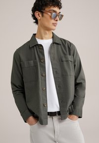 Olijfgroene button-up shirt met twee borstzakken, lichtgewicht stof en bruine knopen, gedragen over een wit T-shirt en lichtgekleurde broek.