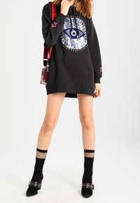 Nainen, jolla on yllään musta ylipitkä hupparimekko, jossa on iso silmäkuvio, verkkosukat, mustat korkokengät ja pieni punainen olkalaukku.