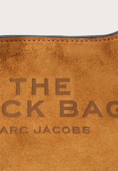 Suede brown bag detail with stitched edge and embossed text "THE BACK BAG MARC JACOBS" on textured surface.
