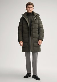 Cappotto imbottito verde scuro, trapuntato, con colletto alto, zip frontale e chiusura a bottoni, indossato sopra un dolcevita aderente e pantaloni grigi.