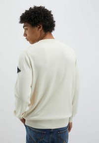 Maglione in cotone crema con polsini e orlo a coste, caratterizzato da una finitura testurizzata e un emblema nero sulla spalla sinistra.