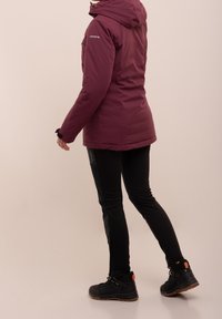 Veste isolante bordeaux avec capuche, dotée de poignets élastiques. Associée à un pantalon noir slim et des bottes noires avec des accents orange.