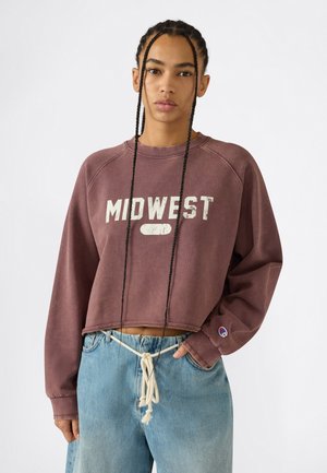 Sweat-shirt court bordeaux avec un graphisme "MIDWEST" ; il comporte des manches raglan et un col côtelé. Associé à un jean en denim bleu clair, ample.