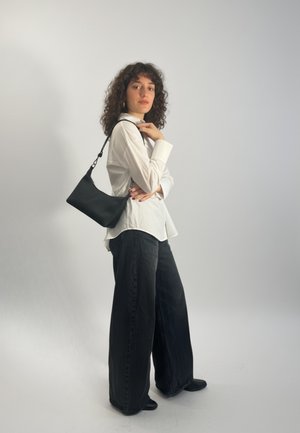 Czarna skórzana torba na ramię z pojedynczym paskiem, gładką fakturą i minimalistycznym designem. Modelka nosi białą koszulę i szerokie czarne spodnie.