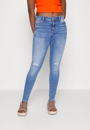 Jeans skinny azul claro com detalhes rasgados nos joelhos, combinados com um top cropped laranja brilhante e sandálias castanhas com tiras.