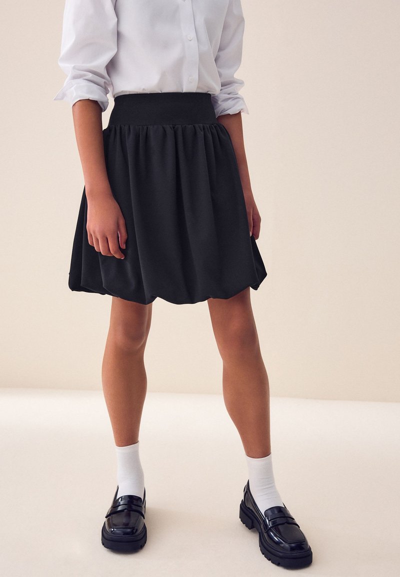 Falda negra con textura fruncida, una cintura alta y una silueta acampanada. Combinada con mocasines negros y calcetines blancos.