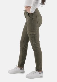 Olivgröna cargobyxor med sidofickor, en figursydd design och avsmalnande anklar, parat med vita sneakers med grå detaljer.
