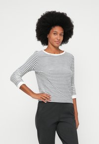 Mulher com cabelo afro natural, vestindo uma camisa de mangas longas cinza e branca com riscas e calças escuras, em pé com uma mão na anca.