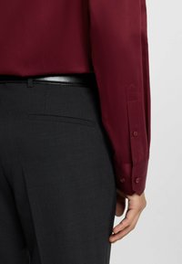 Camicia bordeaux a maniche lunghe con polsini abbottonati, indossata con pantaloni grigi scuri su misura dotati di tasca posteriore e una cintura nera.