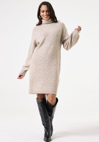 Beige gestricktes Sweatkleid mit hohem Kragen, strukturiertem Muster und langen Ärmeln; kombiniert mit knielangen schwarzen Lederstiefeln.