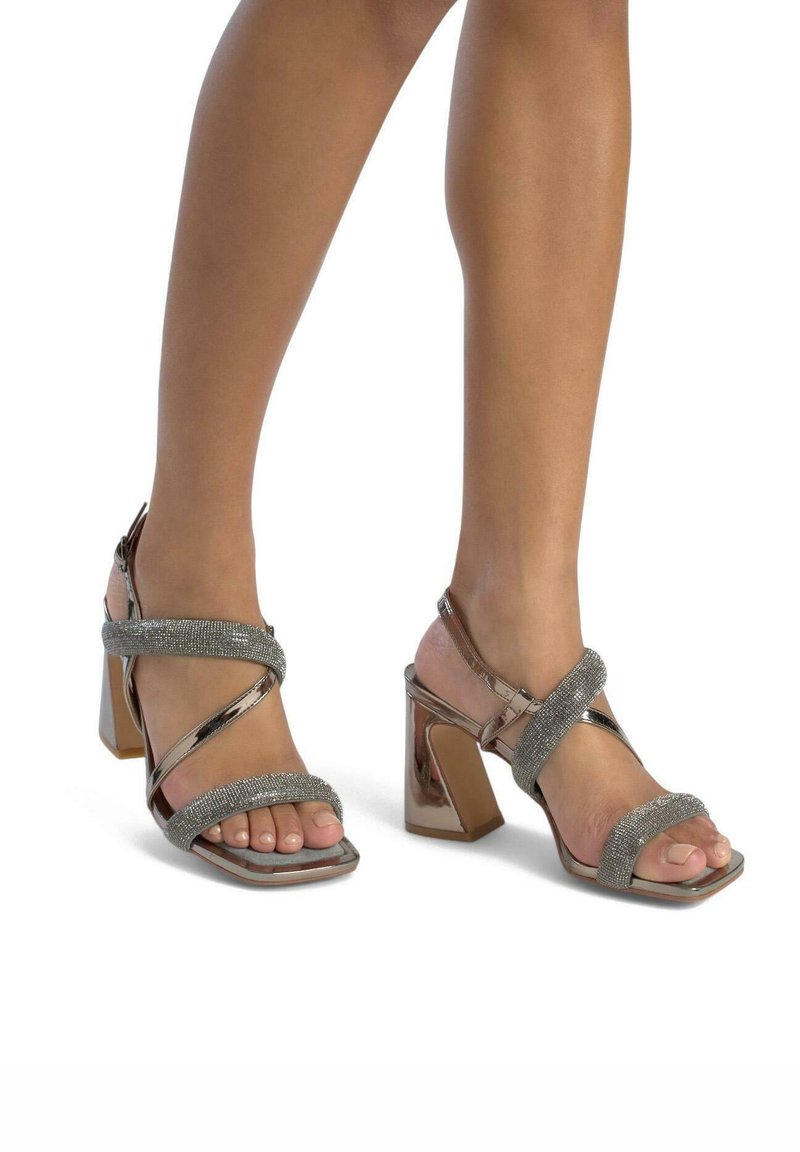 Sandalias de tacón bloque abierto con acabado metálico en oro rosa y tiras entrelazadas decoradas con detalles brillantes. Caja de dedos en forma cuadrada.