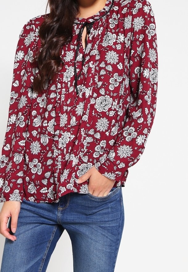 Femme portant un chemisier rouge à manches longues avec des motifs floraux blancs et un nœud noir au niveau du col, associé à un jean bleu.