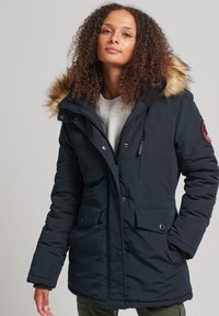 Marineblauwe parka met een capuchon met bontafwerking, vooraan met ritssluiting, drukknopen, meerdere zakken en een gestructureerde stof.