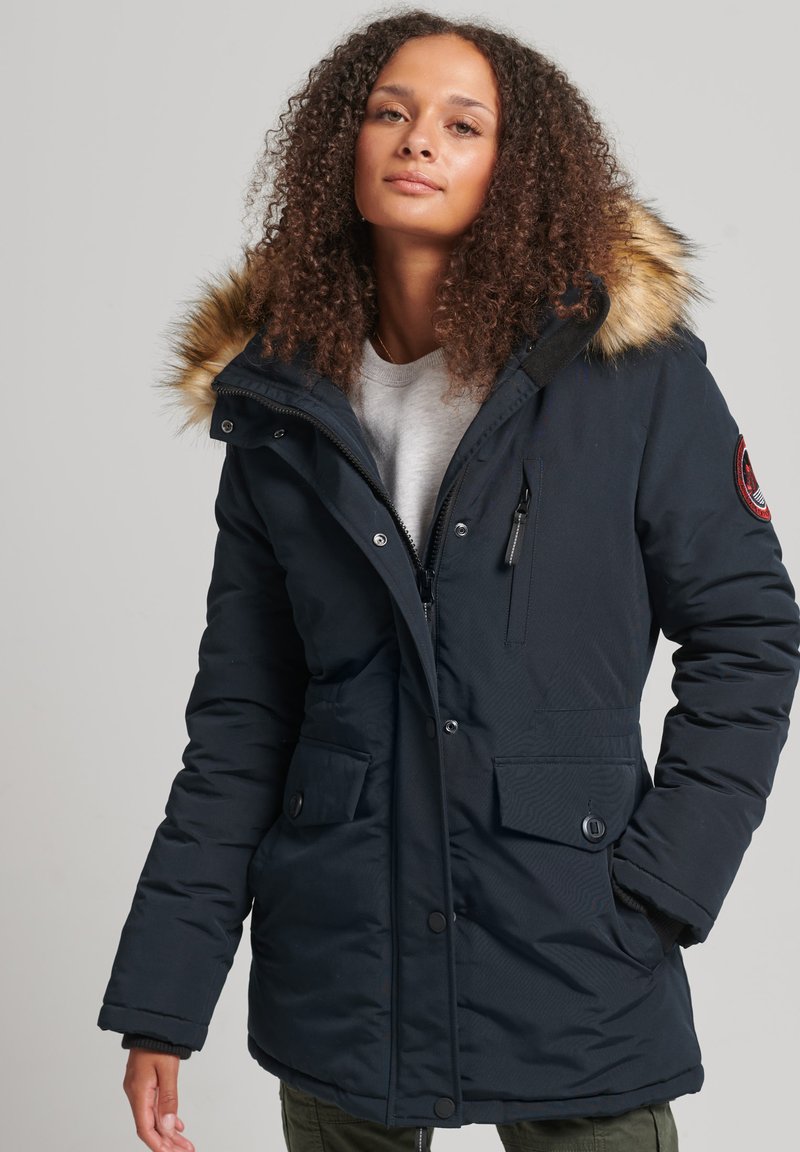 Marineblauwe parka met een capuchon met bontafwerking, vooraan met ritssluiting, drukknopen, meerdere zakken en een gestructureerde stof.