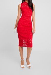Robe en dentelle rouge avec une silhouette ajustée, sans manches et une longueur au genou. Des talons blancs à brides et un petit sac à main blanc sont également présents.