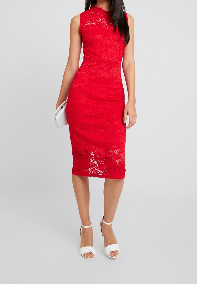 Robe en dentelle rouge avec une silhouette ajustée, sans manches et une longueur au genou. Des talons blancs à brides et un petit sac à main blanc sont également présents.