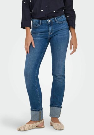 Femme portant un jean bleu à revers, des chaussures plates tissées beiges et une chemise noire à manches longues avec des motifs de cœurs blancs.