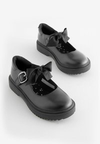 Chaussures en cuir noir avec un bout arrondi, une bride de cheville et un grand détail en nœud. La semelle texturée offre une adhérence ; l'intérieur présente un motif d'étoiles.