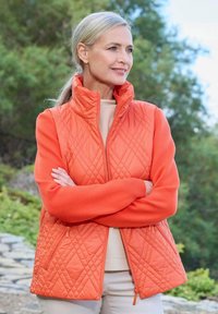 Eine Frau mittleren Alters mit blonden Haaren, die eine orange gesteppte Weste und einen Pullover trägt, steht im Freien mit verschränkten Armen und schaut nach rechts.