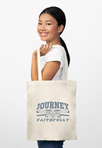 Bolsa de lona en un suave color beige con un diseño gráfico que presenta texto en azul que dice "JOURNEY FAITHFULLY" y acentos de alas.