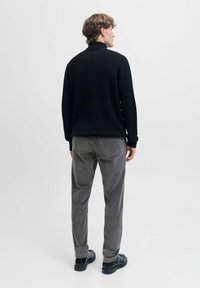Schwarzer gerippter Pullover mit hohem Kragen, kombiniert mit grauen strukturierten Hosen und schwarzen Schuhen, von hinten vor einem neutralen Hintergrund betrachtet.