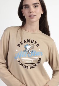 Camisa de manga larga beige con un gráfico de Snoopy usando una gorra, con texto negro y azul que dice "PEANUTS All Star Beagle Playing Since 1950."