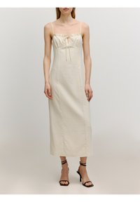 Langes, cremefarbenes Kleid mit dünnen Trägern, gerafftem Vorderteil mit einer Schleife und gerader Silhouette; getragen mit schwarzen, hohen Sandalen.