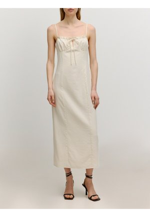 Lange, crème kleurige jurk met dunne straps, geplooide voorkant met een strik en een rechte silhouette; gedragen met zwarte hakken sandaaltjes.