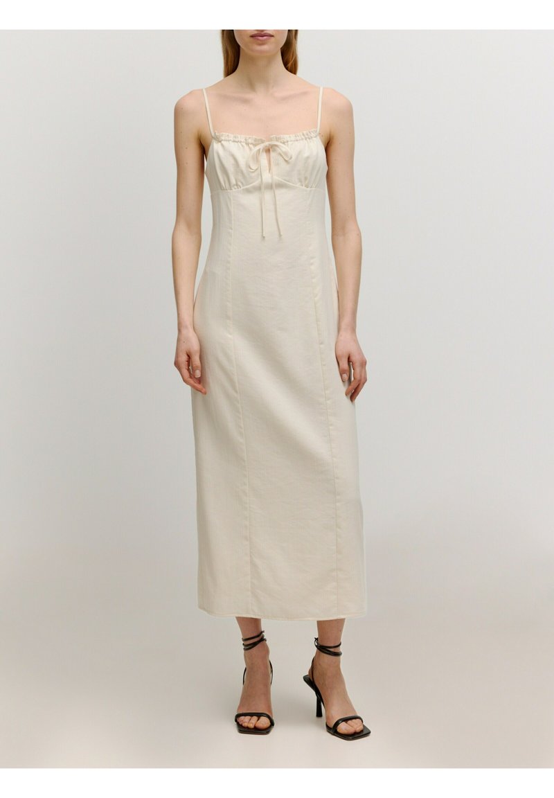 Langes, cremefarbenes Kleid mit dünnen Trägern, gerafftem Vorderteil mit einer Schleife und gerader Silhouette; getragen mit schwarzen, hohen Sandalen.