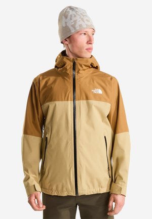 Unge mand iført to-tonet tan og brun North Face jakke og beige mønstret beanie, der kigger til siden mod hvid baggrund.