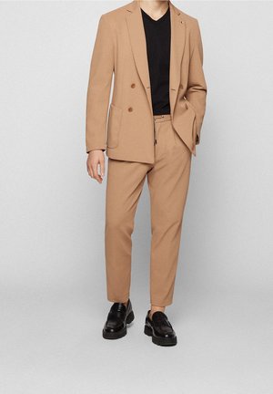 Man draagt een beige dubbelrijkostuum met bijpassende broek, zwart V-halshemd en zwarte loafers, staand tegen een effen achtergrond.