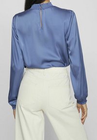 Satinbluse in Blau mit langen Ärmeln und gerafften Bündchen, versehen mit einem Knopfverschluss am Hals und kombiniert mit hoch taillierten, hellcremefarbenen Hosen.