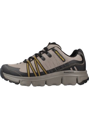Zapatilla atlética beige con detalles en negro y amarillo, parte superior de malla, suela gruesa y diseño texturizado. Presenta un sistema de cordones para un ajuste seguro.