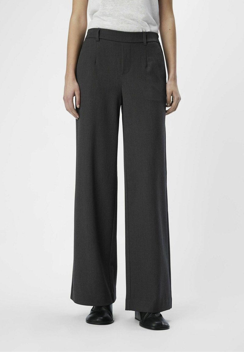 Personne portant un pantalon taille haute à jambes larges gris foncé avec passants pour ceinture et des chaussures noires sur un fond clair uni.