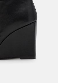 Chaussure à talon compensé noir avec une tige en cuir texturé et un design géométrique. Présente une forme lisse et courbée avec une base solide pour un bon maintien.