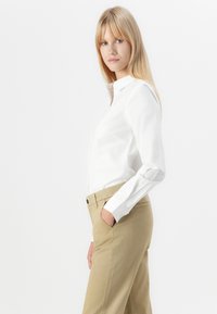 Fehér, galléros, hosszú ujjú ing, rendezett szabással, bézs nadrággal párosítva. Sima anyag és klasszikus dizájn.