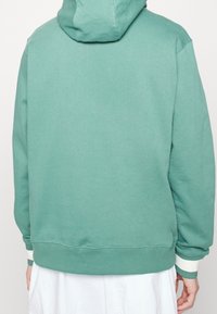 Mikina s kapucí z měkké zelené látky s volným střihem. Má žebrované manžety a lem v kontrastní krémové barvě. Jednoduchý design, bez viditelných log.
