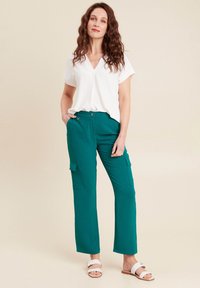Pantaloni cargo color teal con due tasche laterali, abbinati a una maglietta bianca a scollo a V. L'outfit è completato da sandali bianchi. Texture liscia.