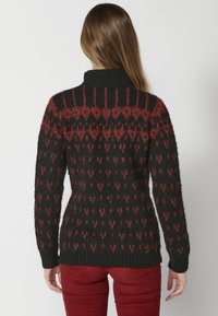 Pull en tricot avec un col montant, présentant un motif géométrique noir et rouge foncé. La texture semble douce et pelucheuse, avec des manches ajustées.
