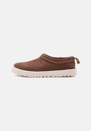 Καφέ παπούτσι τύπου slip-on με πλεκτή διακόσμηση, λευκή χοντρή σόλα και το λογότυπο UGG στο πλάι, εμφανίζεται σε προφίλ σε απλό φόντο.