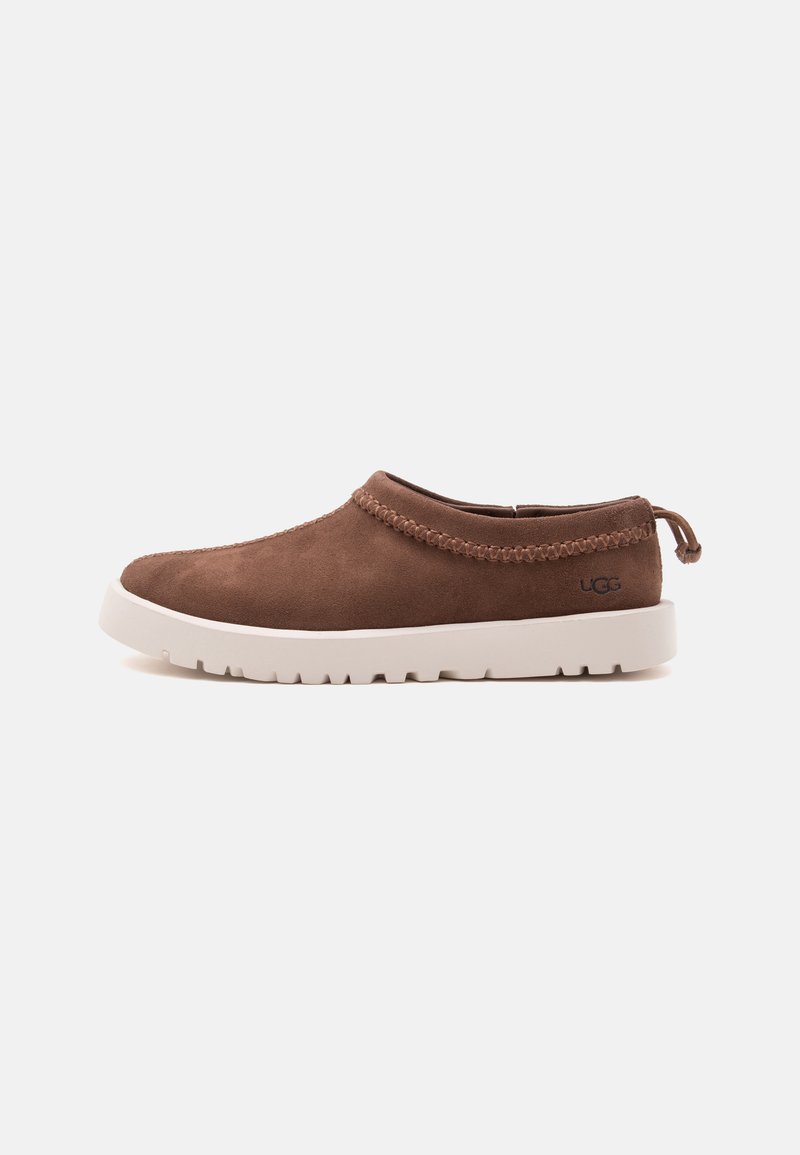 Καφέ παπούτσι τύπου slip-on με πλεκτή διακόσμηση, λευκή χοντρή σόλα και το λογότυπο UGG στο πλάι, εμφανίζεται σε προφίλ σε απλό φόντο.