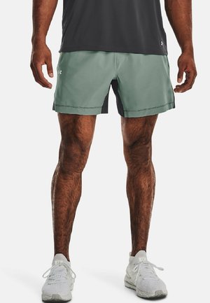Man wearing light green athletic shorts, black undershorts, white sneakers, and a dark gray shirt, standing with legs slightly apart.