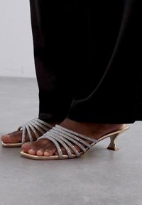 Sandales à lanières argentées avec des accents scintillants, un talon courbé en or et un design à bout ouvert. La texture est lisse et brillante.