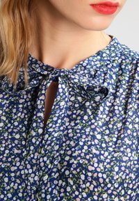 Navybluse mit floralem Muster und zartem Halsband, gestaltet mit kleinen rosa und weißen Blumen, grünen Akzenten und leichtem Stoffdesign.