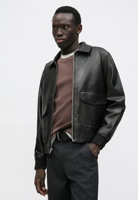 Casaco de couro preto com fecho de zíper na frente, gola e dois bolsos frontais, usado sobre um suéter bordô texturizado com gola redonda.