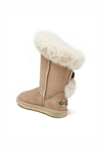 Beige suede boot with a cream fur cuff, round toe, flat sole, and a decorative tag on the side. Features a plush pom-pom at the back.