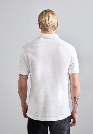 Lyshåret mand set bagfra, iført en hvid kortærmet polo og mørke bukser, med synlige tatoveringer på begge underarme.